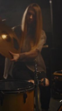 Uzun saçlı beyaz kadın müzisyenin perküsyon enstrümanlarını ayarlayarak canlı performansa hazırlandığı dikey bir sahne.
