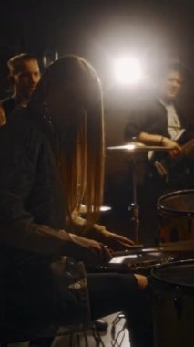Uzun saçlı genç bayan davulcunun, loş ışık stüdyosunda gitarda eşlik eden erkek grup arkadaşlarıyla birlikte perküsyon enstrümanları çalarken orta boy dikey çekimi.