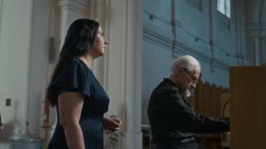 Yaşlı bir org çalgıcısıyla müzik provası yapan kadın vokalist yanında durmuş kilisedeki müzik standından notalar okuyor ve şarkı söylüyor.