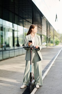 Kafkasyalı genç bir iş kadınının elinde akıllı bir telefonla dışarıda e-scooterıyla dikey çekim yaparken görüntüsü.