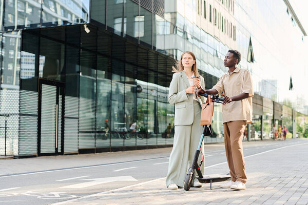 Широкий снимок белой женщины, держащей чашку кофе, и ее чернокожего коллеги-мужчины с электронным скутером, болтающего на улице