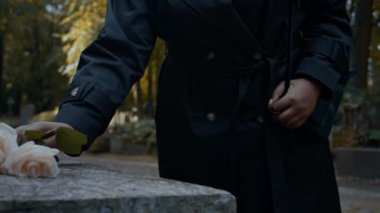 Siyah trençkotlu, tanınmayan bir kadının mezarlığı ziyaret ederken bir çift güzel beyaz gülü mezar taşına koyarkenki yakın çekimi.