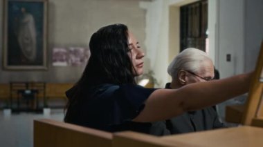 Esmer kadın şarkıcı, gri saçlı yaşlı erkek orgcunun önündeki müzik standına müzik notaları koyuyor, sonra da katedralde notalar okuyor.