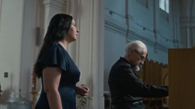 Yaşlı bir org çalgıcısıyla müzik provası yapan kadın vokalist yanında durmuş kilisedeki müzik standından notalar okuyor ve şarkı söylüyor.