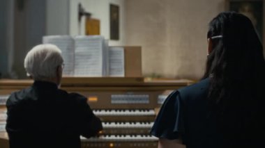 Esmer kadının müzik notalarına bakarken enstrüman çalan kıdemli orgcu eşliğinde müzik parçalarını seslendirdiği arka plan görüntüsü
