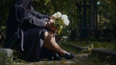 Siyah elbiseli, yas tutan bir kadının mezarlıkta oturup taş yola gül attığı bir sahne.