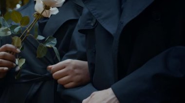 Mezarlıktaki anma töreninde el ele duran ve gülleri tutan siyah ceketli çiftin yakın çekimleri.
