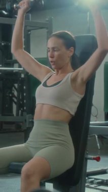 Spor salonunda ağırlık çalışırken genç bir kadının dambılları yukarı kaldırışının dikey çekimi.