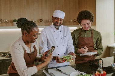 Afro-Amerikan kadının şef salatasının fotoğrafını çekip blog 'a koyduğu yüksek açılı bir fotoğraf. Gururlu şef salata tabağı tutuyor ve gülümsüyor. Kıvırcık tavuklu aşçı yemek tariflerini yazarken.