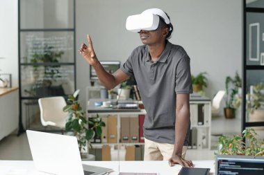Üzerinde elektronik cihazlar olan çalışma masasının yanında dururken VR kulaklıklı Afro-Amerikalı bir adam oyun oynuyor.