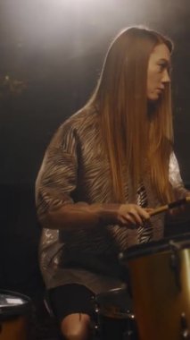 Genç bayan müzisyenin loş bir konser sahnesinde prova sırasında davul çaldığı dikey çekim.