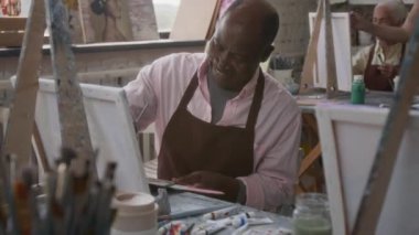 Resim dersinde önlük giymiş gülümseyen siyah bir adam, arka planda yaşlı bir kadın ve genç bir öğretmen.