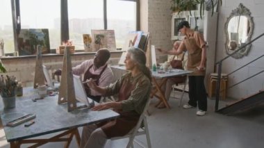 Sanat dersine katılan bir grup son sınıf öğrencisi, stüdyoda masalarda otururken tuvallere resim çiziyorlar, kadın öğretmen de resimli kadınlara yardım ediyor.