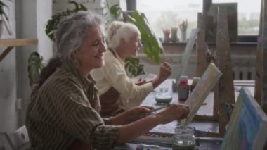 Kafkasyalı son sınıf öğrencisinin tuvale balmumu boya kalemleriyle çiçek boyadığı, siyah adamın resim dersinde onun yanında çalıştığı, arka planda başka bir kadının olduğu görüntüyü büyüt.