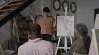 Üç son sınıf öğrencisi stüdyoda resim sehpasında sunulan soyut resimlerden bahsedip duruyor.