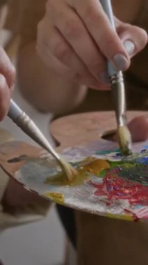 Bilinmeyen yaşlı kadın ve bayan öğretmenin elleri, tahta palette guaj rengiyle resim atölyesindeki fırçaları karıştırırken dikey yakın plan görüntüsü.