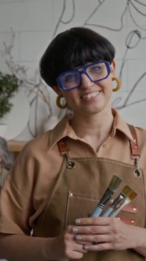 Kısa saçlı esmer kadın ressamın dikey portresi. Önlüklü, kameraya poz veren, elinde tebessümle resim stüdyosunda üç fırça tutan genç kadın.