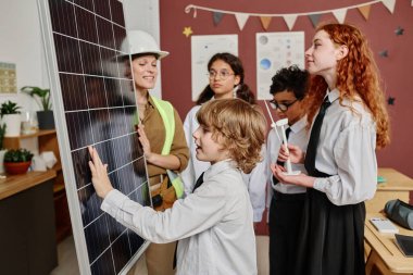 Sınıf arkadaşları kariyer günü boyunca kadın mühendisleri dinlerken beyaz çocuk güneş panelinin yüzeyine dokunuyor.