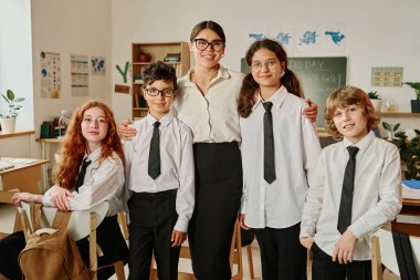 Sınıfta durup kameraya poz veren neşeli bayan öğretmen ve genç kız ve erkek çocukların orta uzunlukta toplu portresi.