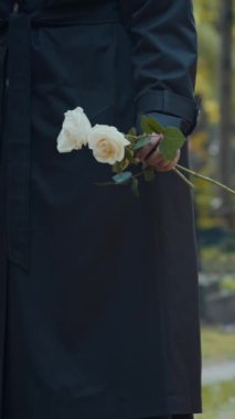 Mezarlıkta dururken elinde bir çift gül tutan, siyah ceketli, tanınmayan bir kadının dikey yakın çekimi.