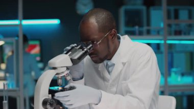 Laboratuvarda çalışırken mikroskop altında numuneleri inceleyen tek kullanımlık eldivenler ve laboratuvar önlüğü giyen odaklanmış siyah erkek bilim adamı.