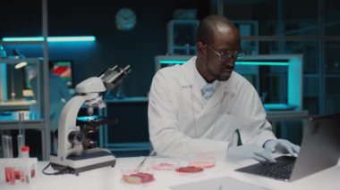 Eldivenli ve laboratuvar önlüklü siyah erkek gıda bilimcisi laboratuvarda çalışırken mikroskop altında kültür ve daktilo örneklerini inceliyor.