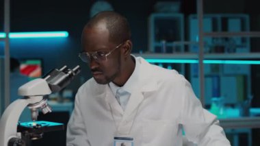 Steril eldiven ve laboratuvar önlüğü giymiş Afro-Amerikan yemek bilimcilerinin mikroskop altında kültürlü et örneklerini incelerken ve araştırma sırasında dizüstü bilgisayarda yazarken aşağı eğilmeleri.