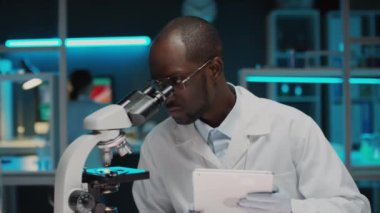 Laboratuvarda moleküler analiz yaparken mikroskoptan bakıp dijital tablet kullanan laboratuvar önlüklü ve eldivenli Afrikalı Amerikalı bilim adamı.