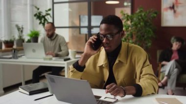 Modern ofiste iş ortağıyla telefonda konuşan Afro-Amerikalı genç erkek yönetici, arka planda iki kişi
