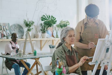 Genç çizim öğretmeni, son sınıf kız öğrencisiyle işbirliği yapıyor. Afrikalı Amerikalı erkek öğrenci arka planda sanatının tadını çıkarırken tuvale resim yapıyorlar.