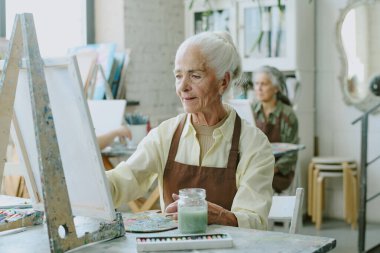 Çalışma masasında resim sehpası, tuval ve çok sayıda boyayla oturan emekli kadın, resim çiziyor ve işleme odaklanıyor.