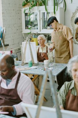 Genç yetişkin resim öğretmeninin arka planda yeni çizim teknikleri öğrenmeye çalışan son sınıf öğrencisini kontrol edişinin dikey çekimi.