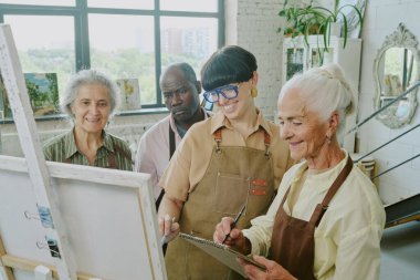 Genç yetişkin öğretmeni ve melez arkadaşları onun resmine bakarken kıdemli kadın ressamların gülümsediği yüksek açılı bir kare.