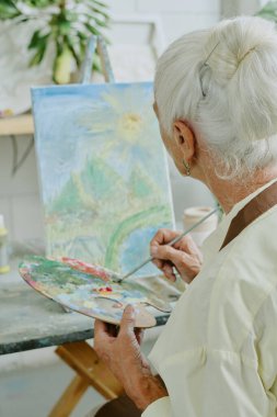 Arka planda, tanınmayan yaşlı bir kadının, ahşap palette yağlı boya karıştırırken tuvalin manzarasına hayran olduğu görüntüler var.