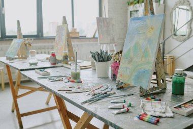 Old wooden tables with acrylic paints, canvases and easels in studio clipart