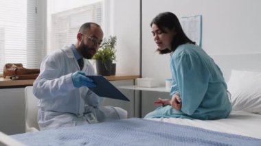Hasta önlüklü kız hastane koğuşunda yatıyor ve ameliyat öncesi konsültasyon sırasında erkek doktorla plastik cerrahi planını tartışıyor.