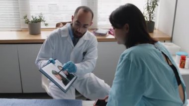 Estetik cerrah, genç bayan hastaya ameliyat öncesi ve sonrası görüntü karşılaştırması yapıyor. Klinikte muayene sırasında dikkatlice dinliyor ve burnuna dokunuyor.