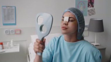 Burnu bandajlı genç bir bayan hasta, ameliyat önlüğü ve şapkası takıyor, yüzünü el aynası ile inceliyor ve sonra klinikte burun ameliyatı sonrası kameraya poz veriyor.