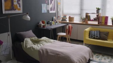 Rahat minimalist yatak odasının içinde tek yatak, masa ve sandalye, sarı kanepe, resimlerle süslenmiş gri duvarlar ve panjurlarla süslenmiş pencereler.