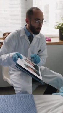 Beyaz önlüklü ve eldivenli doktorun dikey fotoğrafı genç bayan hastada olası yüz değişikliklerini gösteriyor ve hastane koğuşunda onunla yapılacak estetik ameliyatları tartışıyor.