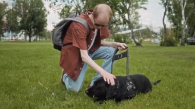 Görme engelli bir adam köpek tasması tutuyor ve parkta çimenlerde uzanmış kara labrador çiğniyor.