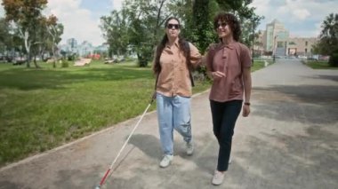 Birlikte parkta yürürken bastonuyla kız arkadaşının omzuna sarılmış görme engelli genç bir kadın.