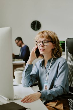 Gözlük takan genç bir ofis çalışanının bilgisayar ekranındaki verilere bakıp telefonla konuştuğu dikey bir fotoğraf.