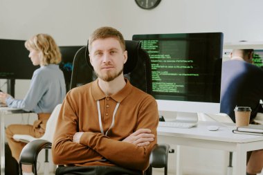 Modern bilgisayarlarla donatılmış, iş yerinde kollarını kavuşturmuş genç beyaz programcının orta boy fotoğrafı.