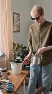 Oturma odasındaki ahşap masada çiçek sulayan görme engelli bir adamın görüntüsüne dikey yakınlaşma