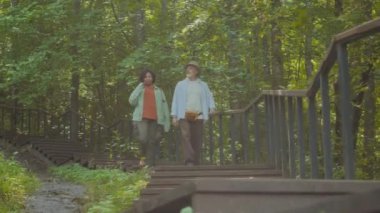 İki yetişkin yolcunun ormanda yürürken ahşap merdivenlerden aşağı inişi alçak açıdan görülüyor.