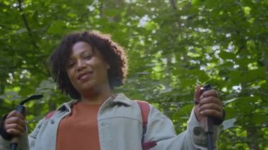 Ormanda yürürken kameraya gülümseyen yürüyüş direkleri kullanan çok ırklı yetişkin bir kadının düşük açılı portresi.