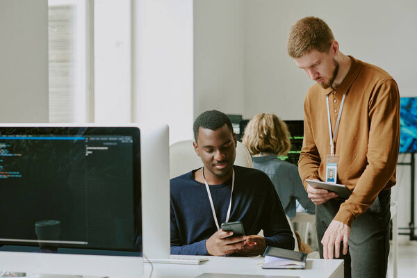Two young ethnically diverse male specialists working together in modern IT company office checking data on digital tablet and smartphone