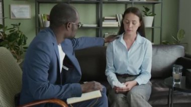 Siyahi erkek danışman, terapi seansında onu rahatlatırken sinirli beyaz kadın müşterinin omzuna dokunuyor.