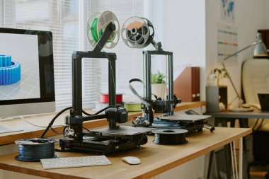 Dual 3D printers operating on wooden desk beside computer displaying object model. Office environment featuring various tech gadgets and decor pieces clipart